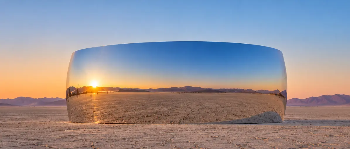 The curved stainless-steel ring seen from the front, its mirror surface reflecting the setting sun, mountains, and cracked desert floor